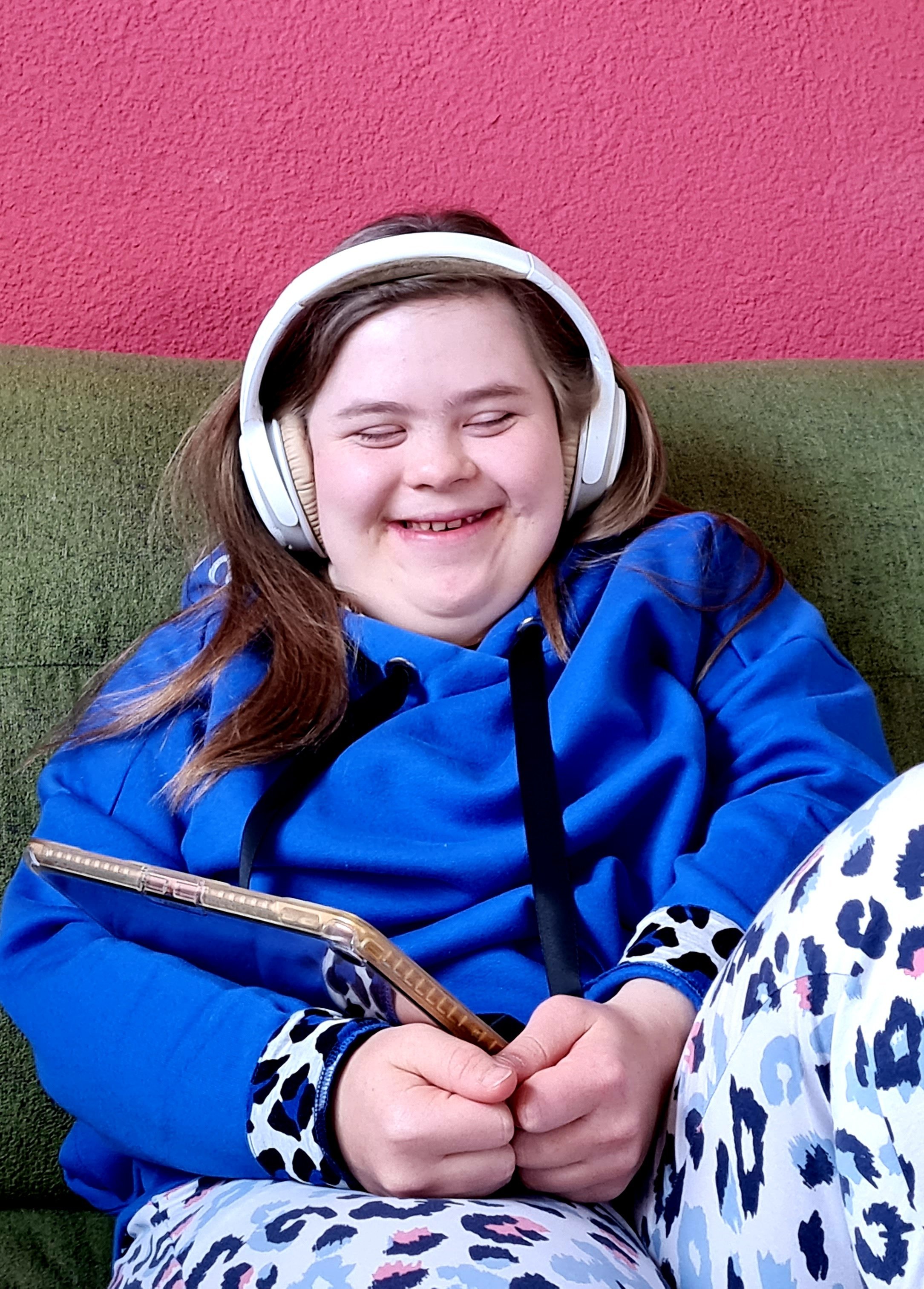 Podcast in Leichter Sprache - Eine junge Frau mit weißen Kopfhörern sitz auf einer grünen Couch. Sie hat ein Tablet in der Hand. Die Augen sind geschlossen und sie lacht fröhlich.